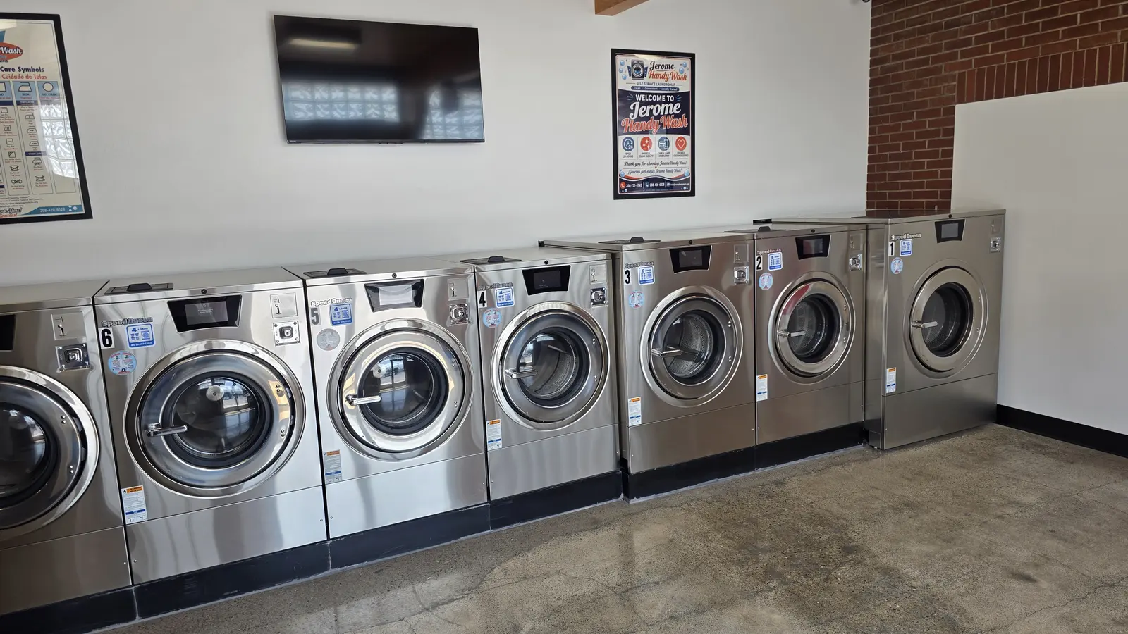 A row of numbered stainless Speed Queen front load washers below a wall mounted television at Jerome Handy Wash